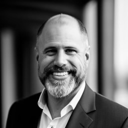 Black and white photo of a smiling man with a bald head and a thick beard He is wearing a suit jacket over a collared shirt and is standing in front of a window The background is blurred drawing focus to his face evoking the calm presence of Zach Small therapist and counselor